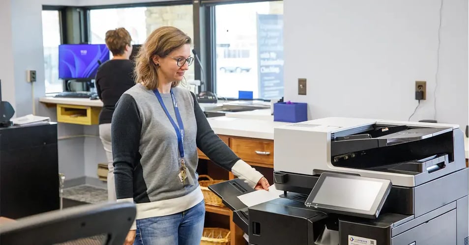 An office professional retrieving a document from a modern multifunction printer, illustrating efficient printer fleet management and device consolidation.