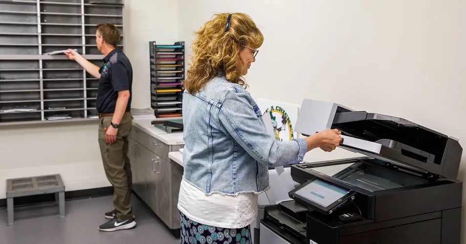 Employee scanning documents at a multifunction copier while a coworker sorts mail showing multi-location print management in action.