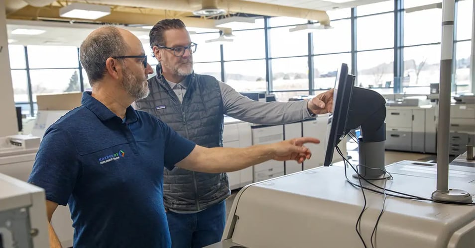 Two production print professionals review a printer’s control panel in a commercial print facility surrounded by production printers.