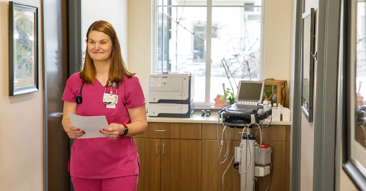A healthcare professional in pink scrubs holds a printed document while standing in a clinic hallway, with a multifunction printer and medical equipment visible in the background.