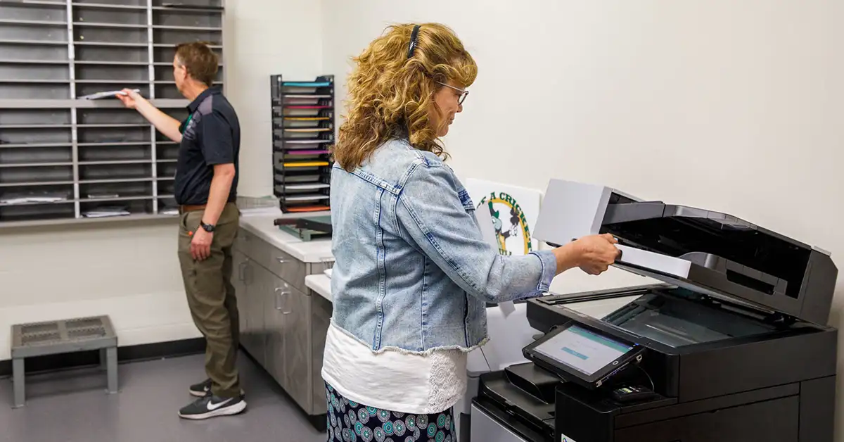 Employee scanning documents at a multifunction copier while a coworker sorts mail showing multi-location print management in action.