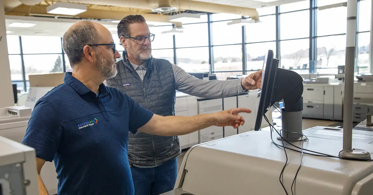 Two production print professionals review a printer’s control panel in a commercial print facility surrounded by production printers.